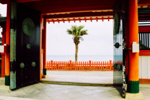 Framed view of a red shrine gate opening to a bright seascape, with a green palm tree and Takachiho-inspired atmosphere from Miyazaki Japan.